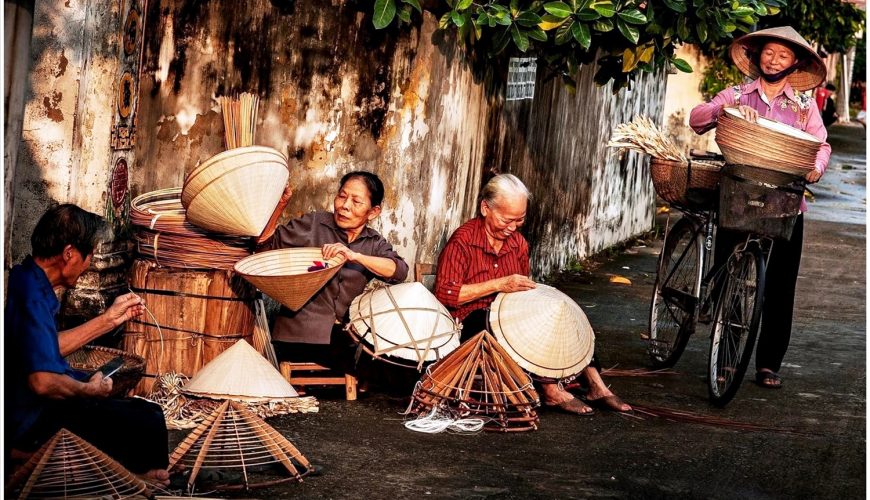 người phụ nữ lớn tuổi đang ngồi làm nón trên vỉa hè, phía sau là một người phụ nữ khác đứng với xe đạp.