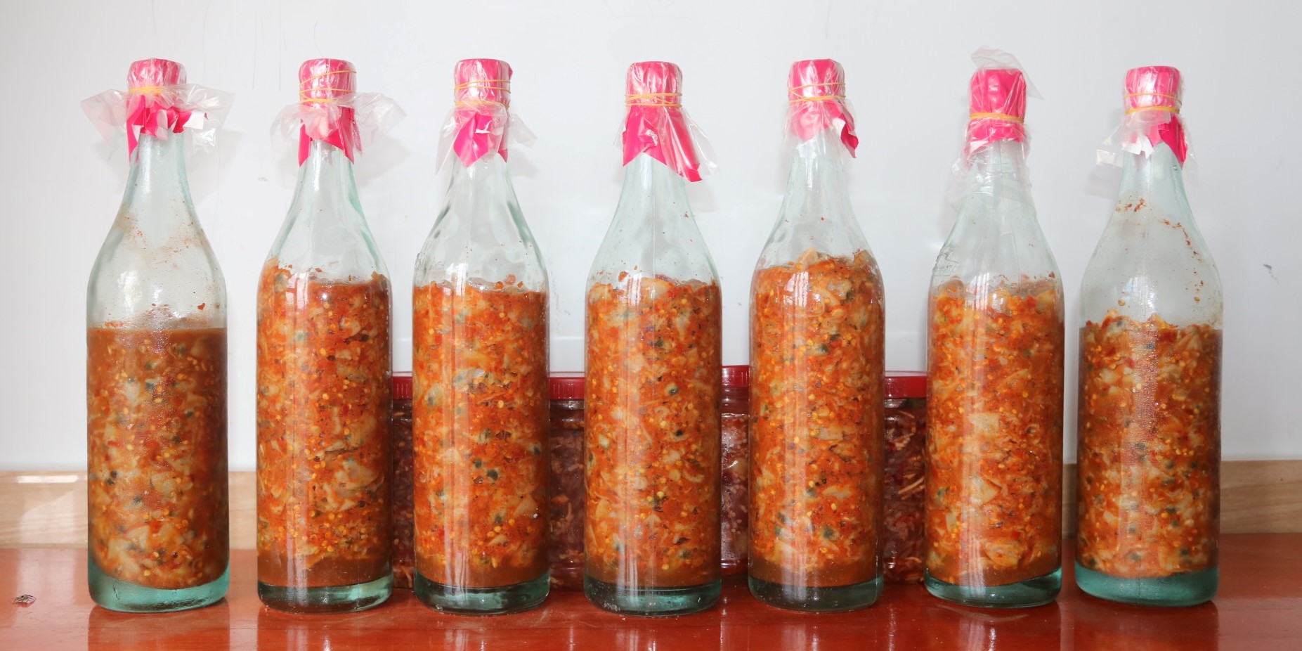 Những chai mắm sò Lăng Cô được đóng trong chai thủy tinh, có màu đỏ cam, đặt trên mặt bàn gỗ.