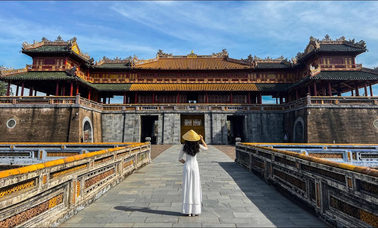 Một người phụ nữ mặc áo dài trắng và nón lá đứng quay lưng, nhìn về phía cổng chính của Quần thể Di tích Cố đô Huế, gợi lên hình ảnh du lịch văn hóa.