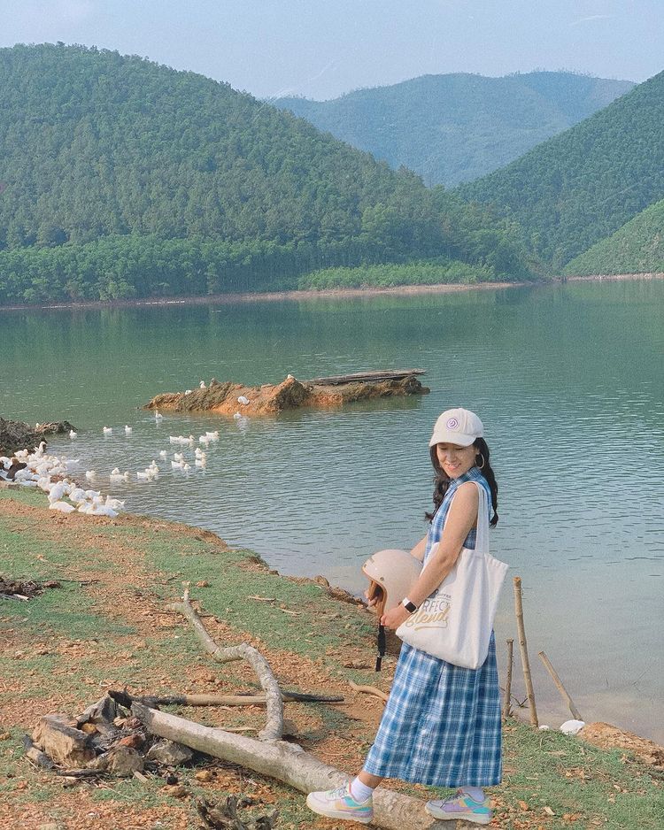 Một cô gái trẻ đội mũ lưỡi trai và cầm mũ bảo hiểm đứng bên bờ Hồ Khe Ngang.