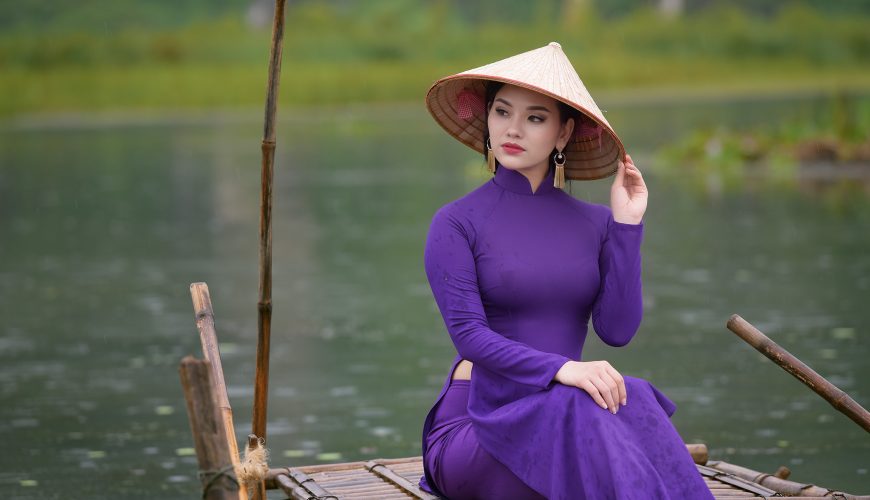 Một cô gái Huế duyên dáng mặc áo dài tím truyền thống, đội nón lá, đứng bên bờ sông Hương.
