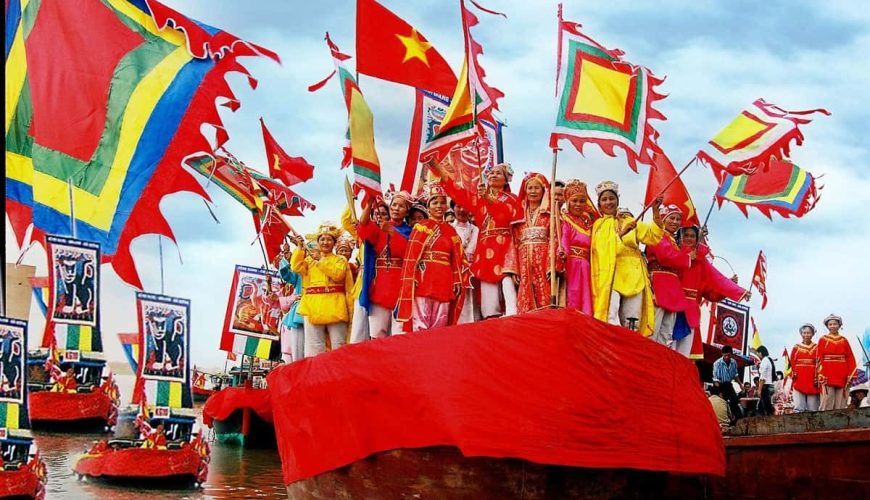 Hình ảnh đoàn người mặc trang phục truyền thống, cầm cờ lọng, cờ phướn nhiều màu sắc, đứng trên thuyền lớn màu đỏ trong Lễ hội Cầu Ngư ở Huế.