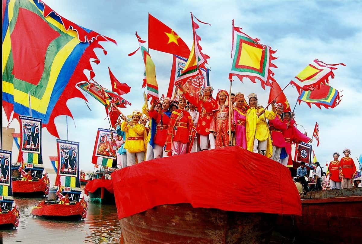 Hình ảnh đoàn người mặc trang phục truyền thống, cầm cờ lọng, cờ phướn nhiều màu sắc, đứng trên thuyền lớn màu đỏ trong Lễ hội Cầu Ngư ở Huế.